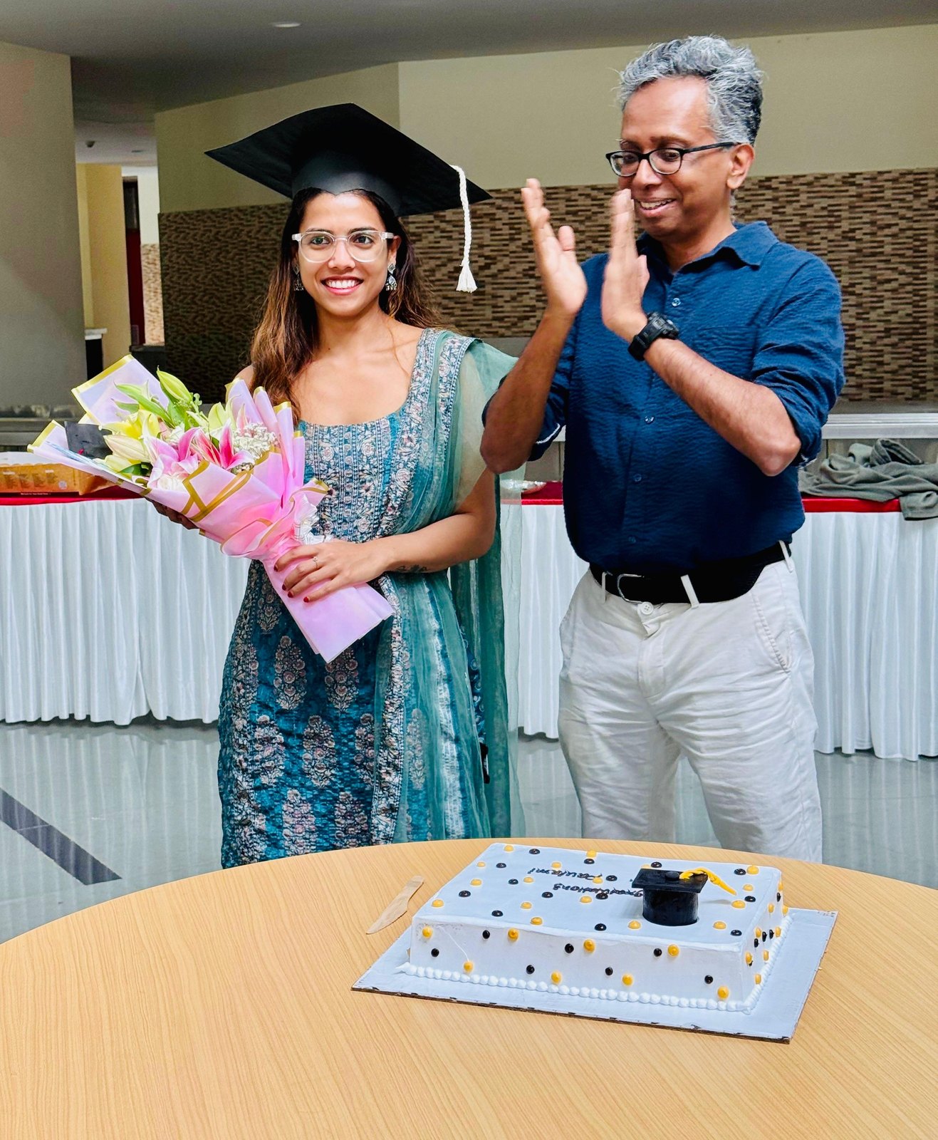 Paulami Dey in her graduation cap holding flowers, with Arvind Ramanathan applauding beside her and a congratulations cake on the table